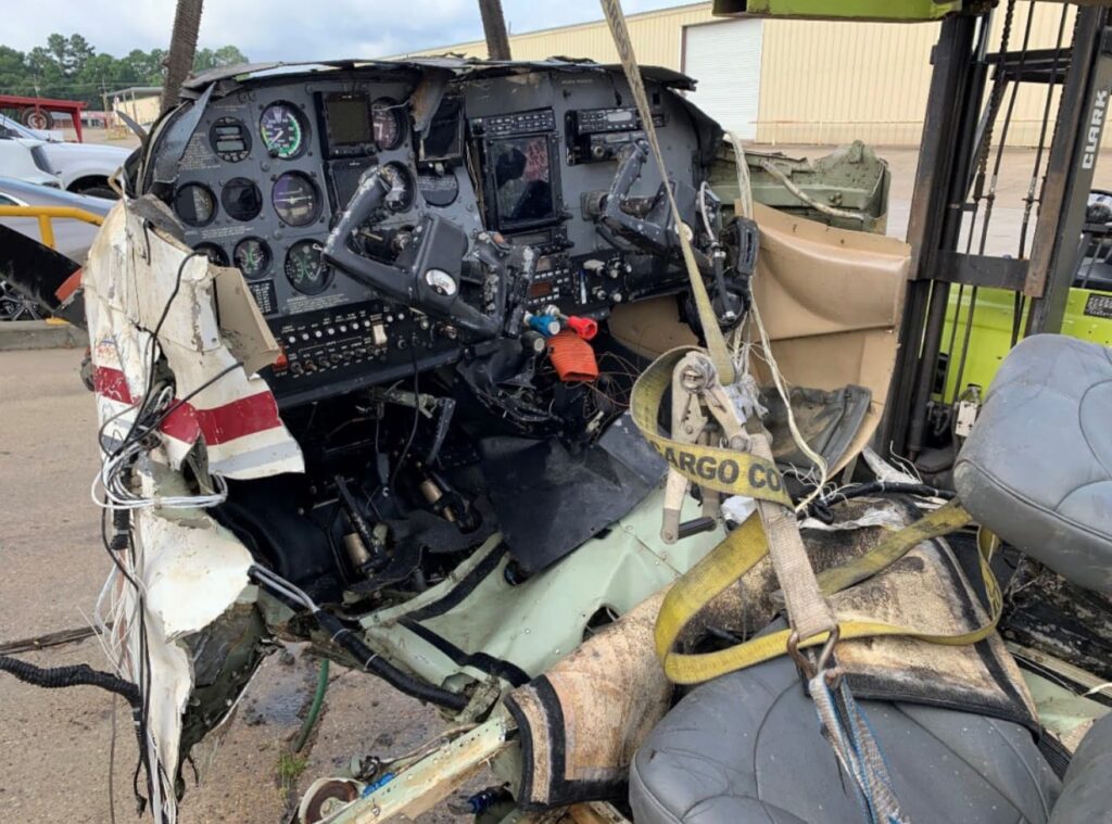 Interior view of a wrecked Cessna 182T cockpit. The instrument panel is crushed and deformed, with broken yokes and exposed wiring. The left side fuselage is torn, and the cabin seats are displaced and damaged. Cargo straps secure the wreckage for transport.